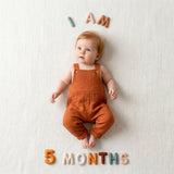 Baby in rust muslin overalls on a cream woven blanket with Neutral Felt Milestone Numbers in earth tones spelling "I AM 5 MONTHS."