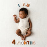 Sleeping baby in a cream linen romper on a soft woven blanket with Neutral Felt Milestone Numbers spelling "I AM 4 MONTHS."