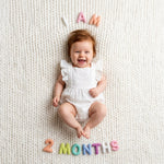 Pastel Felt Milestone Numbers spelling "I AM 2 MONTHS" arranged around a laughing baby on a cream knit blanket.