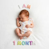 Pastel Felt Milestone Numbers spelling "I AM 1 MONTH" around a sleeping newborn in a pastel rainbow striped romper.
