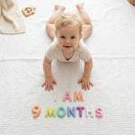 Smiling baby doing tummy time on a white muslin blanket with Pastel Felt Milestone Numbers spelling "I AM 9 MONTHS" and a wooden binoculars teether nearby.
