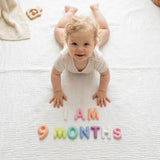 Smiling baby doing tummy time on a white muslin blanket with Pastel Felt Milestone Numbers spelling "I AM 9 MONTHS" and a wooden binoculars teether nearby.