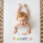 Auburn-haired baby in a rattan crib smiling up at the camera with Pastel Felt Milestone Numbers spelling "I AM 9 MONTHS" beside her hands.