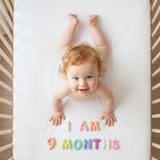 Auburn-haired baby in a rattan crib smiling up at the camera with Pastel Felt Milestone Numbers spelling "I AM 9 MONTHS" beside her hands.