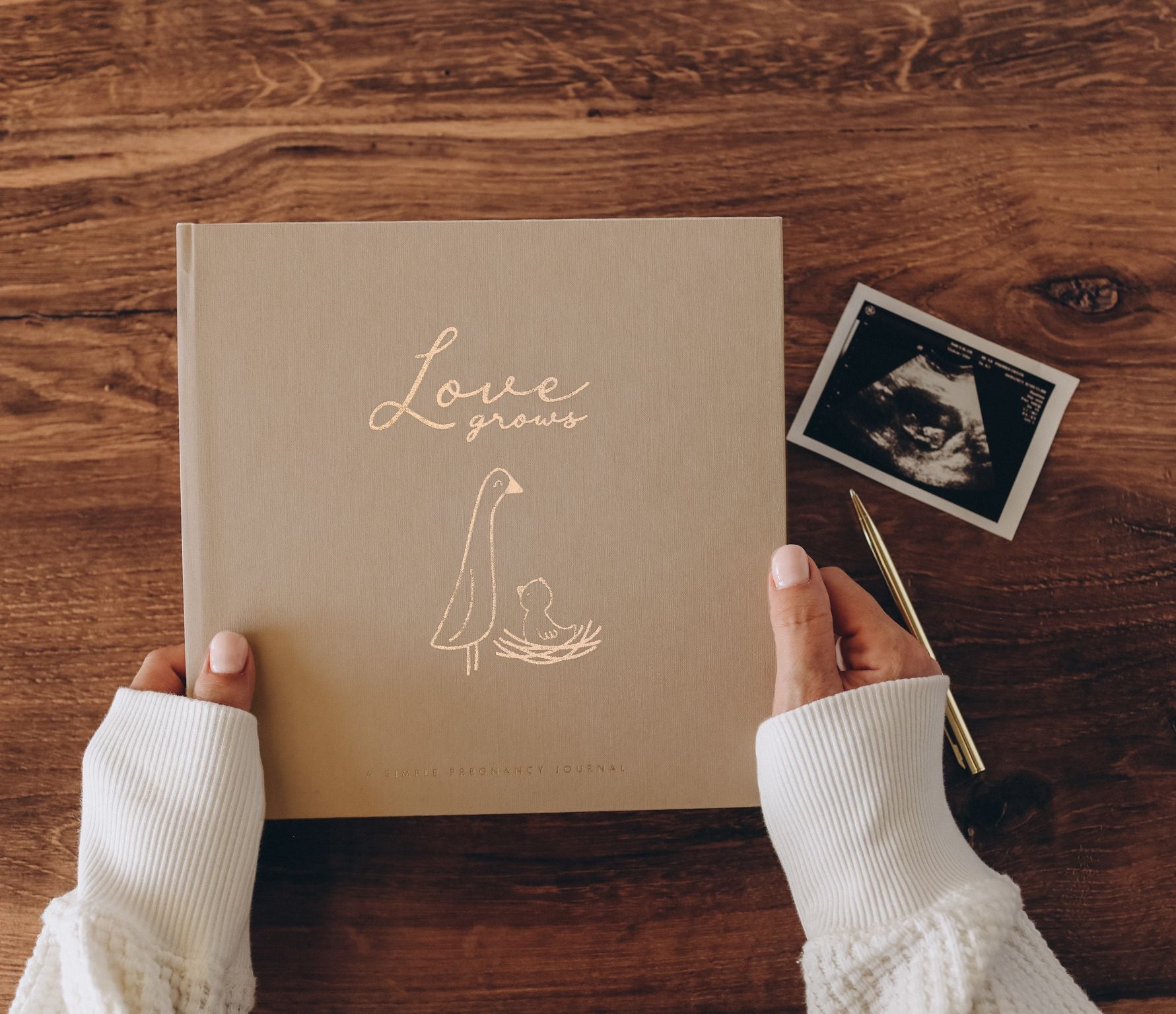 Hands holding the Lucy Darling Pregnancy Journal in Desert Sand on a wood table, with an ultrasound photo and gold pen